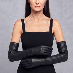 May include: A pair of long black leather gloves featuring a vertical ribbed or quilted texture extending from the wrist up to the forearm. The gloves are smooth at the hand section and have a structured, fitted appearance with a matte finish.