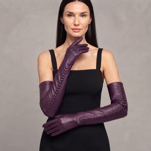 May include: A pair of long, elbow-length purple leather gloves featuring a row of small, matching fabric-covered buttons running vertically along the outer forearm. The gloves have a smooth, matte finish and extend past the elbow, providing full coverage.