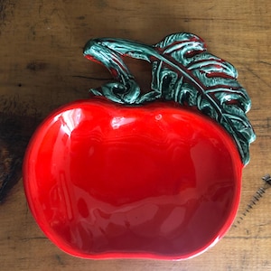 May include: A glossy red ceramic dish shaped like an apple, featuring a prominent dark green stem and a textured leaf extending from the top. The interior of the bowl is smooth and vibrant red, while the exterior leaf detail has dark accents.