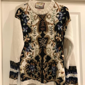 May include: A long-sleeved knit sweater featuring a crew neckline and a complex, multi-colored jacquard pattern in black, cream, tan, and blue. The garment displays intricate botanical-inspired motifs across the front and solid cream sleeves.