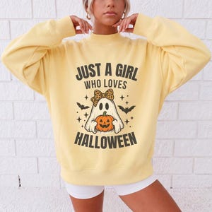 Könnte beinhalten: Ein blassgelbes Langarm-Sweatshirt mit einem Grafikdruck eines Geistes, der eine Schleife mit Leopardenmuster trägt und einen orangefarbenen Kürbis hält. Der Text lautet JUST A GIRL WHO LOVES HALLOWEEN, umgeben von kleinen schwarzen Fledermäusen.