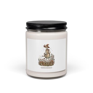 Peut inclure: Une bougie en pot de verre transparent avec un couvercle noir contenant de la cire blanc cassé. L'étiquette montre un squelette portant un chapeau et un tablier, arrosant un jardin de fleurs et de champignons avec un petit oiseau. Texte "GROWING THROUGH IT".
