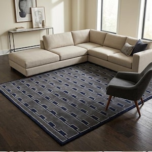 Könnte beinhalten: Ein großer rechteckiger Teppich mit einem sich wiederholenden geometrischen Muster in Anthrazit und Marineblau, der auf einem dunklen Holzboden unter einem beigen Ecksofa und einem dunkelgrauen Sessel mit Holzbeinen liegt.