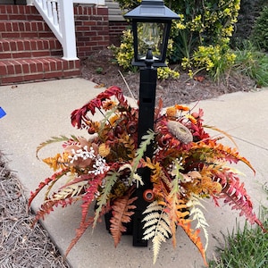 May include: A black solar-powered lamp post centerpiece decorated with an arrangement of artificial autumn foliage, including red, orange, and yellow ferns, sunflowers, and small berries, creating a dense, colorful display around the base.
