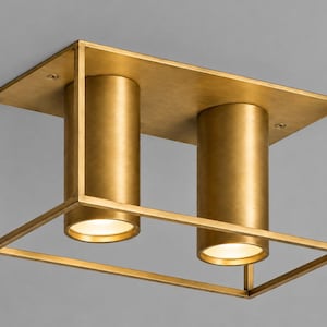 May include: A rectangular ceiling light fixture with a brushed brass finish. The design features a thin metal frame enclosing two cylindrical light sockets that point downward, each emitting a soft, warm glow from the circular base openings.