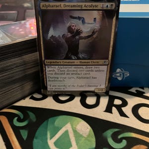 May include: A Magic: The Gathering trading card titled Alpharael, Dreaming Acolyte, featuring a human cleric illustration, text detailing game abilities, and a power/toughness of 2/3, positioned next to a stack of cards and a blue deck box.