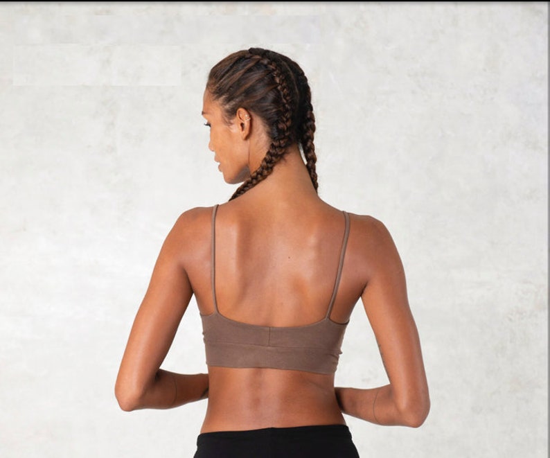 Pode incluir: Suti&atilde; desportivo castanho com al&ccedil;as finas. O suti&atilde; &eacute; usado com cal&ccedil;as pretas. A modelo tem cabelo tran&ccedil;ado. O fundo &eacute; uma parede clara e texturizada.