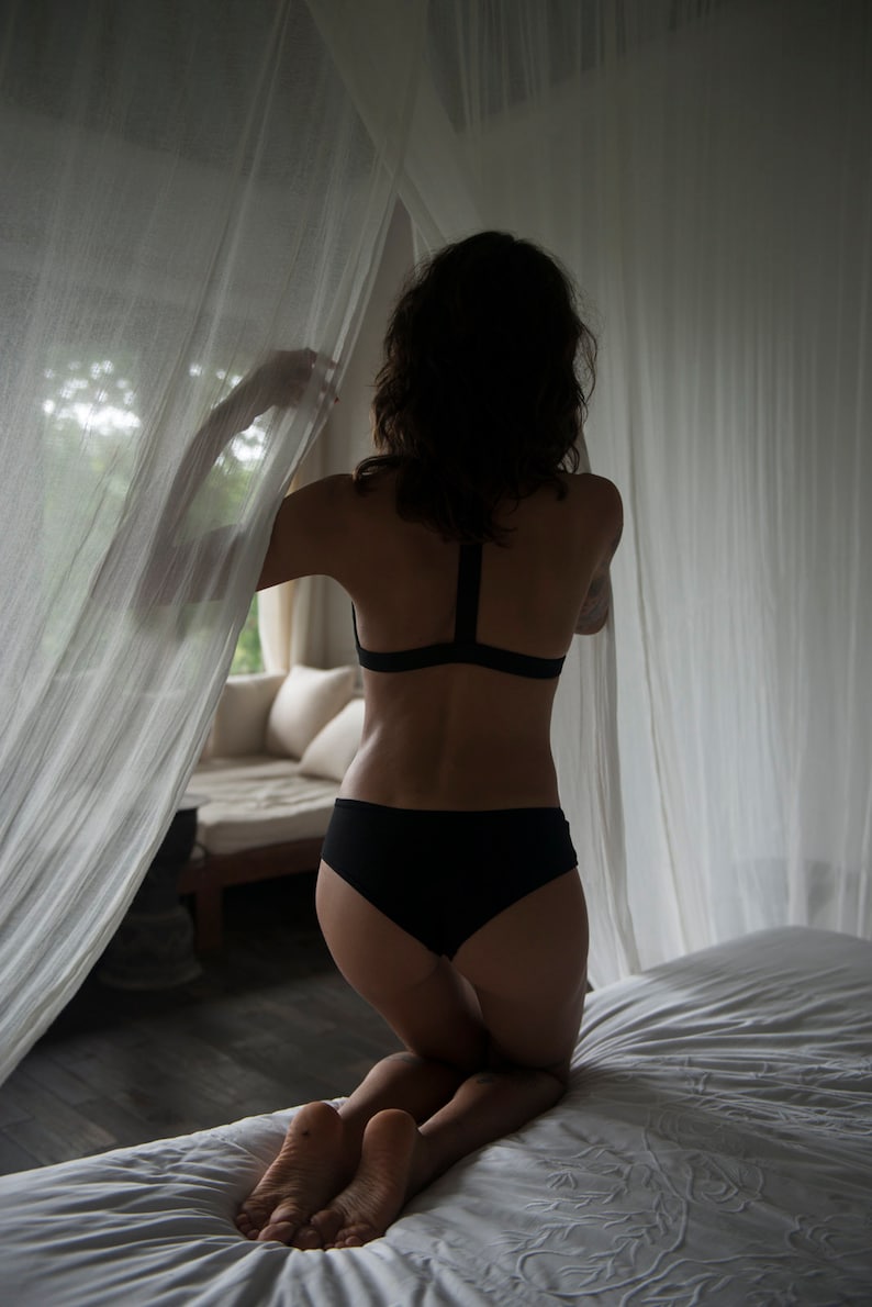 以下が含まれることがあります： 黒いランジェリーを着た人が、白い透き通ったカーテンの前に立って、窓の外を見ています。その人は白いシーツの白いベッドにひざまずいています。