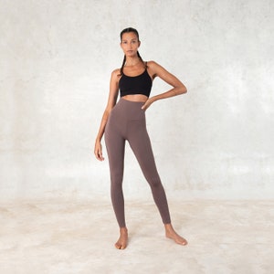Pode incluir: Uma mulher usando um top esportivo preto e leggings marrons de cintura alta. As leggings s&atilde;o de cor s&oacute;lida e parecem ser feitas de um material macio e el&aacute;stico. A roupa &eacute; adequada para ioga, pilates ou outras atividades f&iacute;sicas.
