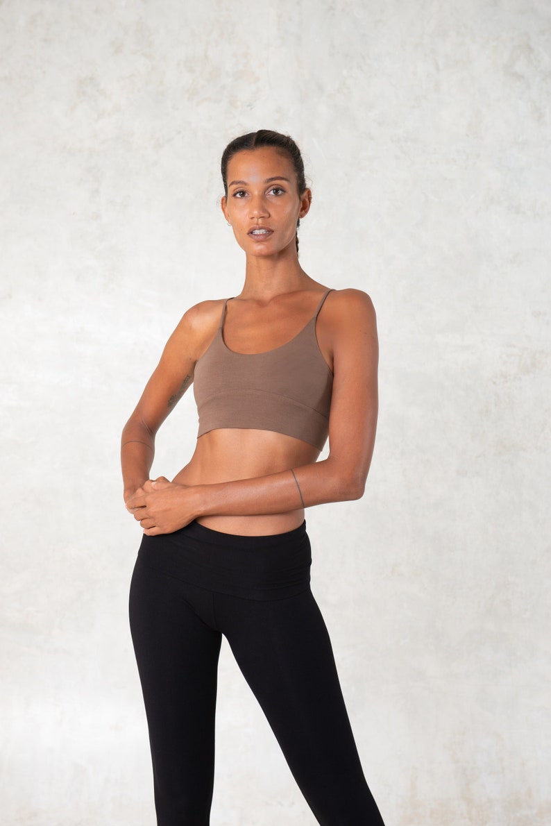 Pode incluir: Uma mulher usando um top esportivo marrom com al&ccedil;as finas e leggings pretas. O top esportivo &eacute; de cor s&oacute;lida e as leggings s&atilde;o de cintura alta. O fundo &eacute; uma parede clara e texturizada.