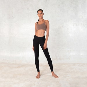 Pode incluir: Uma mulher usando um top esportivo marrom e leggings pretas. O top esportivo tem al&ccedil;as finas e as leggings s&atilde;o justas. O fundo &eacute; uma parede clara e texturizada.