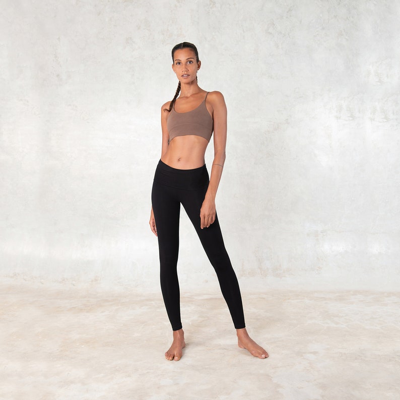 Pode incluir: Uma mulher usando um top esportivo marrom e leggings pretas. O top esportivo tem al&ccedil;as finas e as leggings s&atilde;o justas. O fundo &eacute; uma parede clara e texturizada.