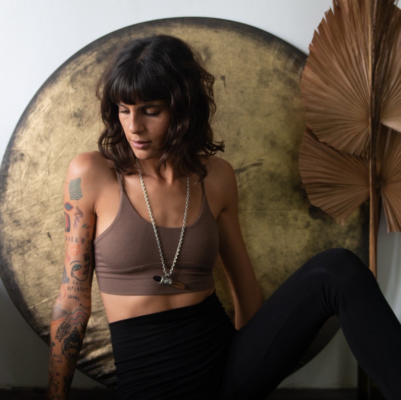 Pode incluir: Uma mulher usando um top esportivo marrom e um colar de corrente de prata com um pingente. Ela est&aacute; usando leggings pretas. Um objeto grande, redondo e dourado est&aacute; atr&aacute;s dela, e uma folha de palmeira seca est&aacute; &agrave; sua direita.
