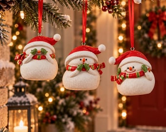 Puede incluir: Tres adornos de muñeco de nieve blancos colgados de cintas rojas. Cada muñeco lleva un gorro de Papá Noel rojo y una bufanda a rayas rojas, verdes y amarillas. Los muñecos tienen narices de zanahoria naranjas y manoplas de color verde.