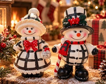 May include: Two snowman figurines featuring black and white plaid patterns. The left figure wears a bonnet with a red bow, while the right figure wears a top hat with holly berries and a red scarf. Both have orange carrot noses and black boots.