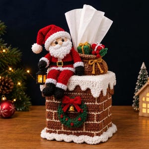 Puede incluir: Una funda de ganchillo para caja de pañuelos con forma de chimenea de ladrillo marrón y borde de nieve blanca. Una figura de Santa Claus se sienta encima con un farol, junto a un saco marrón lleno de regalos. Una corona verde decora el frente.