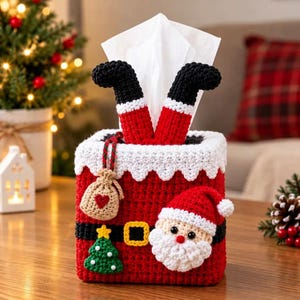 Puede incluir: Una funda de ganchillo para caja de pañuelos en rojo y blanco con forma de chimenea, con las piernas de Santa Claus sobresaliendo por arriba. La funda presenta un cinturón negro con hebilla amarilla, un pequeño saco beige con un corazón rojo, un árbol verde y una cara de Santa Claus.