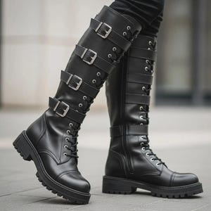 Könnte beinhalten: Ein Paar kniehohe schwarze Stiefel mit Schnürung vorne, silberfarbenen Ösen und vier horizontalen Riemen am Schaft, die jeweils mit einer silberfarbenen Metallschnalle befestigt sind. Die Stiefel haben eine abgerundete Zehenpartie und eine dicke Profilsohle.