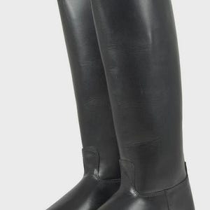 May include: A pair of tall black leather riding boots featuring a smooth finish, a rounded toe, a low block heel, and a structured shaft that extends to the knee. The boots have visible stitching details at the ankle and a clean, minimalist design.