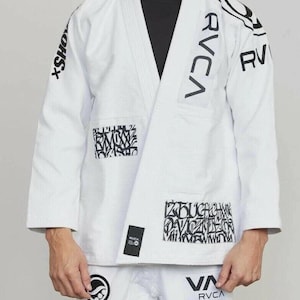 May include: A white martial arts gi jacket featuring black RVCA branding on the chest and shoulders, with black graffiti-style patches on the front panels and a black label near the hem, worn over a plain black shirt with matching white pants.
