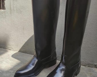 Puede incluir: Un par de botas de montar altas de cuero negro con un acabado liso y pulido, punteras redondeadas y tacón bajo. Las botas presentan una caña estructurada y un forro interior de color tostado. Se ve una tarjeta cuadrada blanca con el texto BOOT BUNKERS.