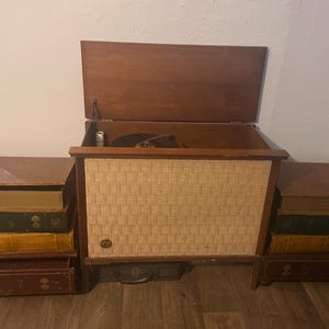 May include: A wooden record player console with a hinged lid and a textured beige speaker grille, flanked by two stacks of decorative storage boxes designed to resemble vintage leather-bound books in shades of brown, green, and yellow.