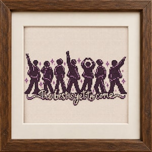 May include: A framed cross-stitch embroidery featuring seven dark purple silhouettes of figures in various poses, accented by small purple stars. Below the figures, the text "The best is yet to come" is stitched in a dark purple, stylized font.