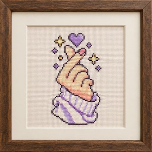 May include: A framed cross-stitch embroidery featuring a hand making a finger heart gesture. The hand emerges from a purple and white ribbed sleeve, surrounded by small purple hearts and yellow and purple four-pointed star sparkles.
