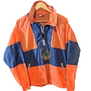 May include: A color-blocked windbreaker jacket featuring bright orange and deep blue panels. The jacket has a high collar, a partial front zipper with a black pull, and two zippered pockets. A circular graphic patch is centered on the chest.