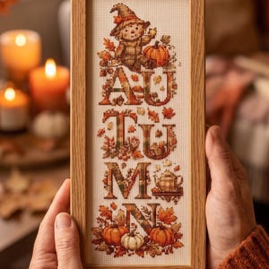 May include: A rectangular wooden frame containing a cross-stitch embroidery on white fabric. The design features the word AUTUMN in large, decorative letters surrounded by fall-themed motifs including a scarecrow, pumpkins, and leaves.
