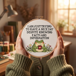 May include: A circular wooden embroidery hoop containing white fabric with a cross-stitch design. The text reads I AM JUST TRYING TO HAVE A NICE DAY DESPITE KNOWING FACTS AND INFORMATION above a green frog holding a red mug with a heart.