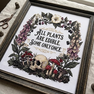 Könnte beinhalten: Ein gerahmtes Kreuzstich-Stickbild auf weißem Stoff mit einem menschlichen Schädel, roten Pilzen und einem Blumenkranz aus Fingerhut und Beeren. Der schwarze Text in der Mitte lautet: All plants are edible. Some only once.