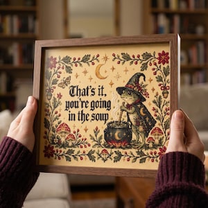 May include: A framed cross-stitch embroidery featuring a frog wearing a witch hat and cape, stirring a black cauldron. The text reads, "That's it, you're going in the soup." The design includes mushrooms, stars, a crescent moon, and a floral border.