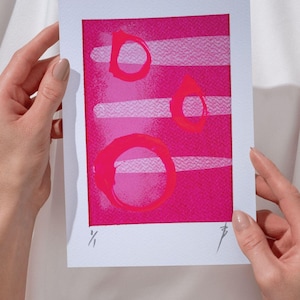 Könnte beinhalten: Abstrakter Kunstdruck mit einem leuchtend rosa Hintergrund, weißen horizontalen Streifen und drei rosa kreisförmigen Formen. Das Kunstwerk wird von Händen gehalten, was seine Größe und Präsentation zeigt. Der Druck ist signiert und nummeriert.