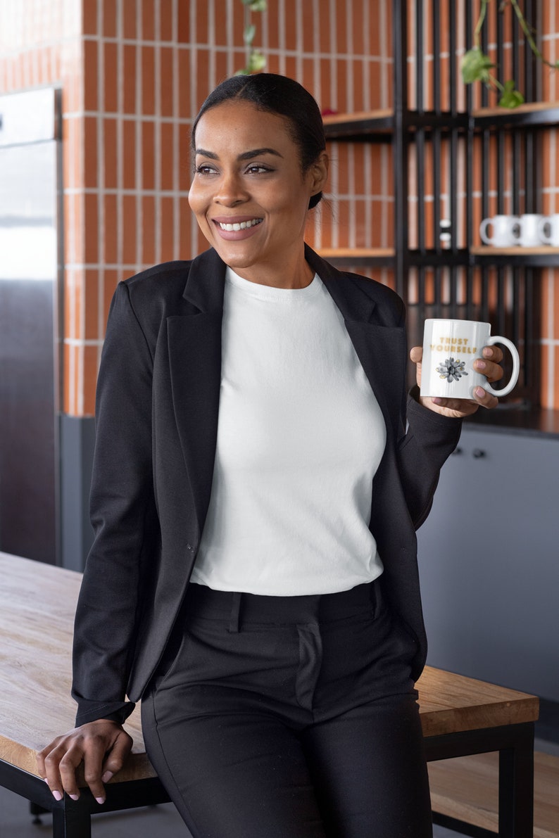 May include: A woman wearing a black blazer and white t-shirt holds a white mug with the text "TRUST YOURSELF" in gold lettering. The mug has a floral design.