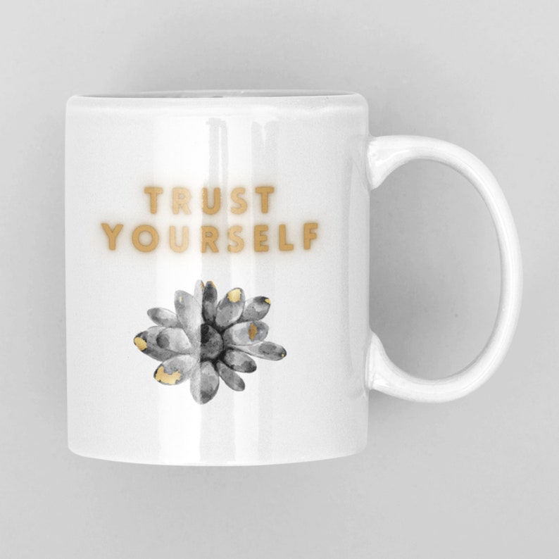 May include: White ceramic mug with a gold inscription that reads "TRUST YOURSELF". A stylized gray and gold succulent is printed on the mug.
