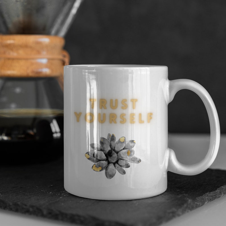 May include: White ceramic mug with a gold design that says "TRUST YOURSELF". The mug has a gray succulent illustration on the front.
