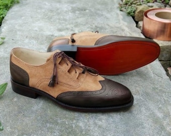 Könnte beinhalten: Ein Paar zweifarbige Oxford-Schuhe mit beigem Wildleder-Obermaterial und dunkelbraunen Lederkappen im Brogue-Stil. Die Schuhe haben dünne braune Schnürsenkel mit Quasten, einen niedrigen Absatz und eine glatte rote Sohle am hinteren Schuh.