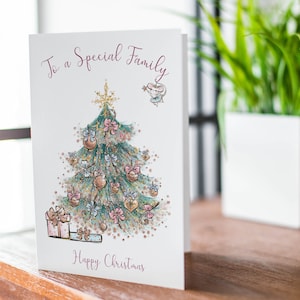 Pu&ograve; includere: Biglietto di Natale con un'illustrazione ad acquerello di un albero di Natale decorato, regali e il testo "To a Special Family" e "Happy Christmas". Il biglietto &egrave; su una superficie di legno.