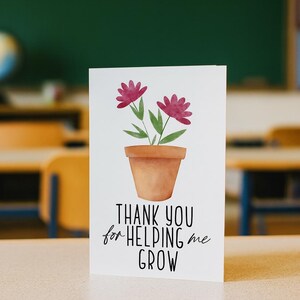 May include: A white card featuring a watercolour illustration of a terracotta flower pot with two pink flowers and green leaves. The text "THANK YOU for HELPING me GROW" is below the illustration. The card is on a desk in a classroom.