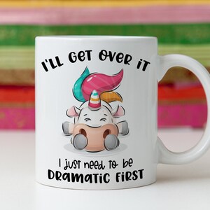 K&ouml;nnte beinhalten: Eine wei&szlig;e Keramiktasse mit einer Cartoon-Einhorn-Illustration. Das Einhorn liegt mit geschlossenen Augen und hat eine rosa, blaue und gelbe M&auml;hne. Der Text auf der Tasse lautet "I'll get over it I just need to be dramatic first."