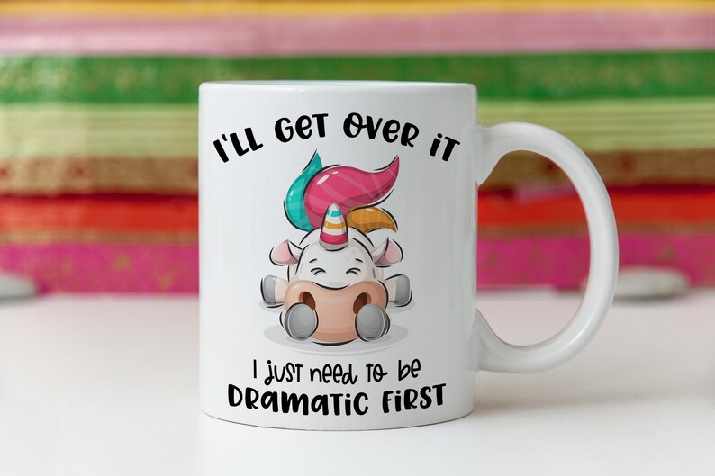 K&ouml;nnte beinhalten: Eine wei&szlig;e Keramiktasse mit einer Cartoon-Einhorn-Illustration. Das Einhorn liegt mit geschlossenen Augen und hat eine rosa, blaue und gelbe M&auml;hne. Der Text auf der Tasse lautet "I'll get over it I just need to be dramatic first."