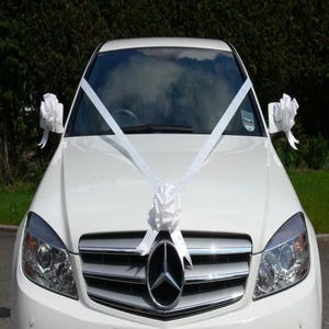 May include: A white car adorned with white ribbons and bows, possibly for a wedding. Ribbons are arranged across the bonnet and on the wing mirrors. The car features a silver grille and a prominent emblem.