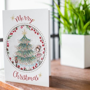 May include: A white Christmas card with a red "Merry Christmas" greeting. The card features a circular illustration of a decorated Christmas tree, a ballerina, and a nutcracker. The illustration is surrounded by a red berry garland.