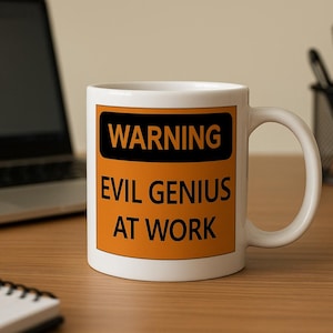 K&ouml;nnte beinhalten: Wei&szlig;e Keramiktasse mit dem Text "WARNING EVIL GENIUS AT WORK" in Schwarz und Orange. Die Tasse steht auf einem Holzschreibtisch neben einem Laptop, einem Notizblock und einem Stifthalter.