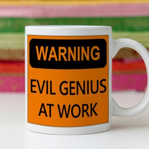 K&ouml;nnte beinhalten: Wei&szlig;e Keramiktasse mit Henkel. Die Tasse hat ein schwarzes und orangefarbenes Warnschild mit der Aufschrift "WARNING EVIL GENIUS AT WORK" in fetten schwarzen Buchstaben. Der Hintergrund ist unscharf.