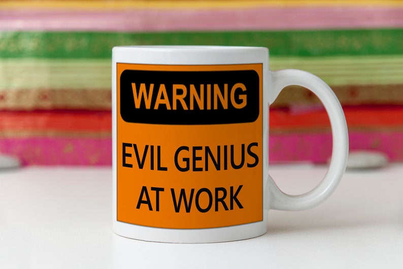 K&ouml;nnte beinhalten: Wei&szlig;e Keramiktasse mit Henkel. Die Tasse hat ein schwarzes und orangefarbenes Warnschild mit der Aufschrift "WARNING EVIL GENIUS AT WORK" in fetten schwarzen Buchstaben. Der Hintergrund ist unscharf.