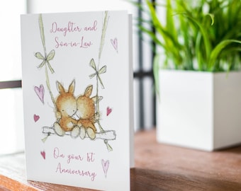 May include: A white card with pink hearts and the text "Daughter and Son-in-Law" and "On your 1st Anniversary". The card features a watercolor illustration of two bunnies on a swing.