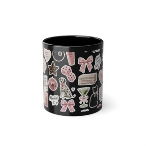Op de afbeelding: Een zwarte keramische mok met een herhalend patroon van diverse stickers in roze, witte en zwarte tinten. De ontwerpen bevatten een dalmatiër, een martiniglas, roze strikken, dobbelstenen, een discobal, boeken, een kat en tekst zoals Manifest It.