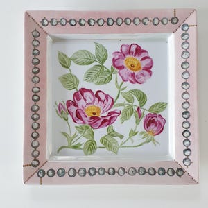 Könnte beinhalten: Ein quadratischer Keramikteller mit einer handgemalten Illustration von rosa Wildrosen mit grünen Blättern in der Mitte. Der breite Rand ist in einem zarten Rosaton gehalten und mit einem wiederkehrenden Muster aus kleinen grauen Kreisen.